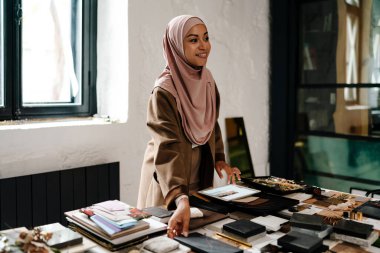 Tesettür giyen neşeli Arap kadın tasarımcı atölyedeki tablet bilgisayarda çalışıyor