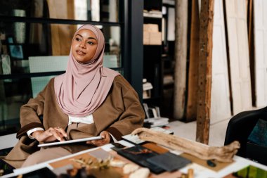 Düşünceli kadın Müslüman tasarımcı tesettür giyiyor atölyede tablet üzerinde çalışıyor