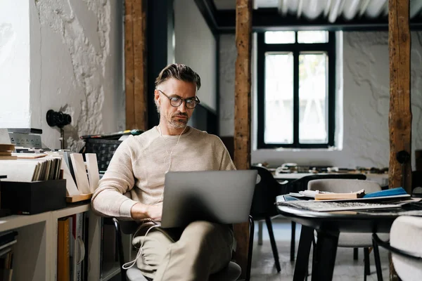 White middle-aged man in earphones using laptop computer while sitting in modern office