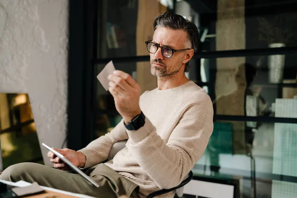 Mature grey man examining samples while working with tablet computer in office