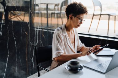 Orta yaşlı iş kadını cep telefonu kullanıyor ve ofiste dizüstü bilgisayarla çalışıyor.