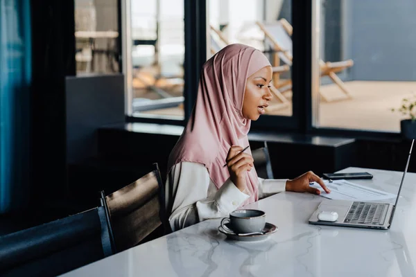Young muslim woman wearing headscarf using laptop and working with documentation in office
