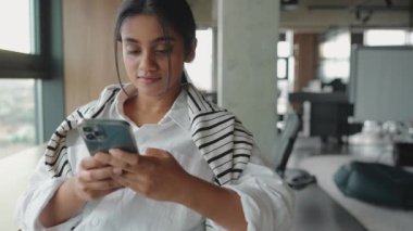 Ofisin penceresinin yanında otururken cep telefonunda yazan neşeli Hintli kadın.