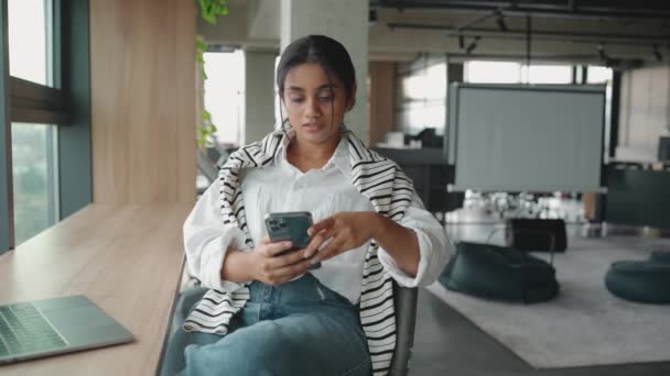 携帯電話を入力し オフィスの窓の近くのノートパソコンで作業するインドの女性 — ストック動画