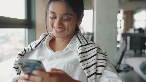 Χαμογελώντας Ινδή Δακτυλογραφεί Στο Τηλέφωνο Ενώ Κάθεται Κοντά Στο Παράθυρο — Αρχείο Βίντεο