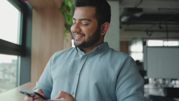 陽気なインド人男性は電話でテキストメッセージを送信し オフィスの窓の近くの側を見て — ストック動画