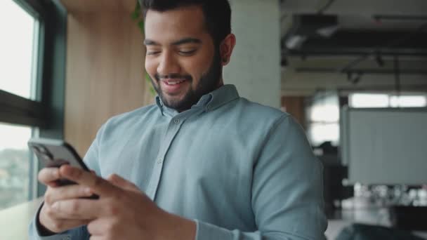 Sorrindo Índio Homem Mensagens Texto Celular Perto Janela Escritório — Vídeo de Stock