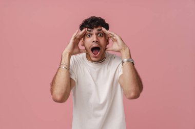 Geniş açık gözleri ve ağzı olan, iki eline dokunan ve kameraya bakan, izole edilmiş pembe arka planda duran genç, yakışıklı ve şık bir adamın portresi.