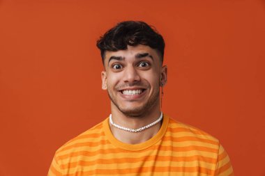 Kolyesi ve tişörtü delinmiş, turuncu arka planda dikilirken kameraya bakan yakışıklı, şık, mutlu bir adamın portresi.