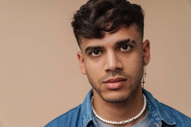 Kolyesi ve kot gömleği olan, yalnız kahverengi arka planda dikilirken kameraya bakan yakışıklı bir adamın portresi.