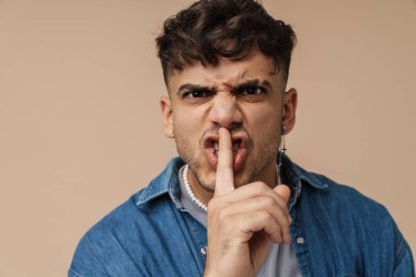 Esmer, suratını buruşturan ve bej arka planda izole edilmiş kameraya sessizlik hareketi yapan genç bir adam.