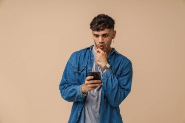 Esmer genç şaşkınlığını dile getiriyor ve bej arka planda izole edilmiş cep telefonu kullanıyor.