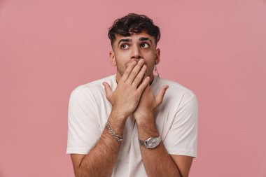 Esmer genç adam pembe arka planda ağzını kapatırken yana bakıyor.
