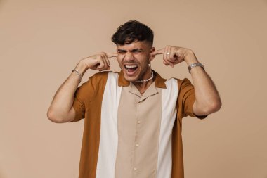 Esmer genç adam, bej arka planda kulaklarını tıkarken çığlık atıyor.