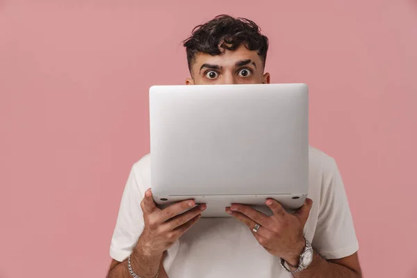 Young handsome curly man peeking from behind a laptop and looking at camera, while standing over isolated pink background