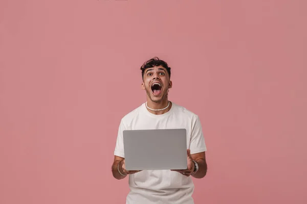 Young handsome enthusiastic man with opened mouth holding laptop and looking upward , while standing over pink isolated background