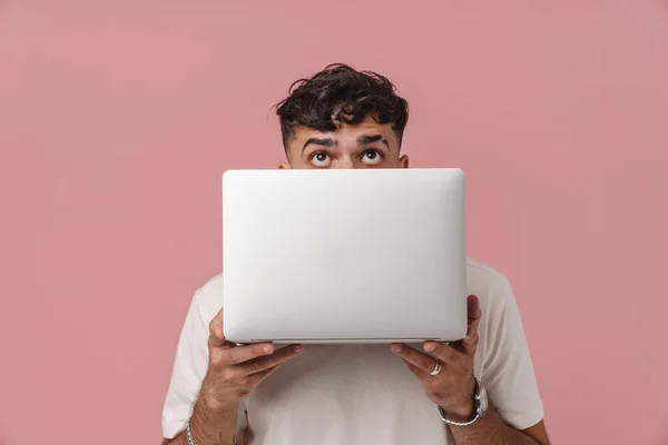 Young handsome curly man peeking from behind a laptop and looking upward, while standing over isolated pink background