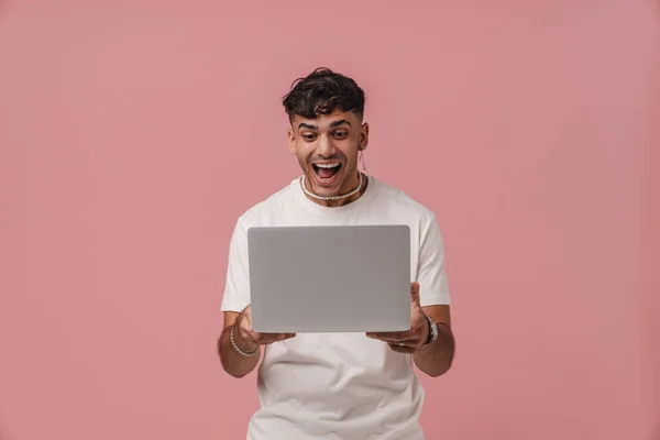 Young handsome enthusiastic man with opened mouth holding laptop and looking on it, while standing over pink isolated background