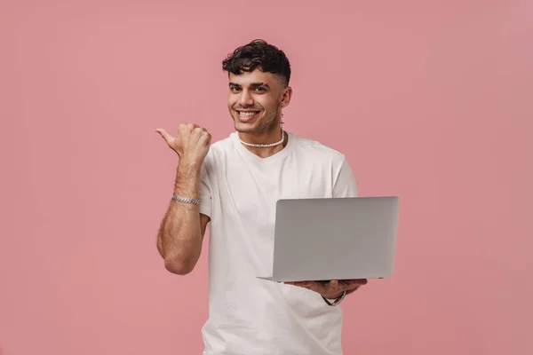 Young handsome enthusiastic man with opened mouth holding laptop, pointing aside with thumb, and looking at camera, while standing over isolated pink background