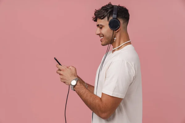 Young hadsome stylish smiling man in headphones with phone standing with his side over pink isolated background