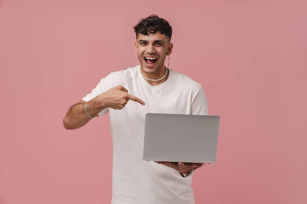 Young handsome enthusiastic man with opened mouth holding laptop and pointing on it, while standing over pink isolated background