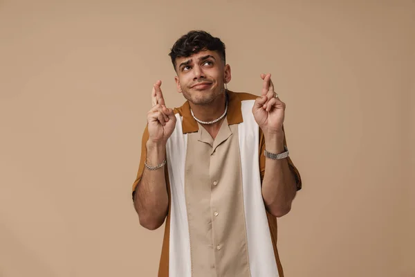 Young handsome stylish hopeful man with pursed lips doing hope gesture with crossed fingers and looking aside , while standing over isolated brown background