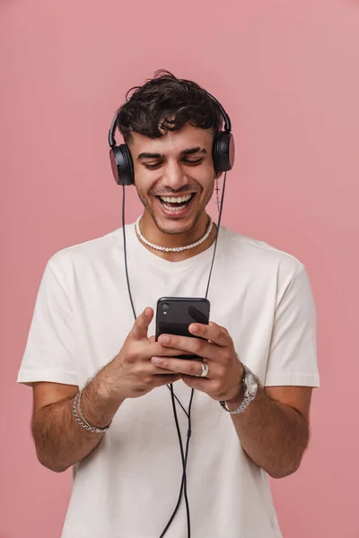 留着耳机的布鲁内特年轻人笑着用粉色背景的手机 — 图库照片