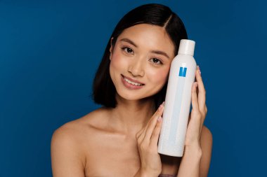 Genç, güzel, gülümseyen Asyalı kadın yüzünün yanında kozmetik ürünler tutuyor ve kameraya bakıyor.