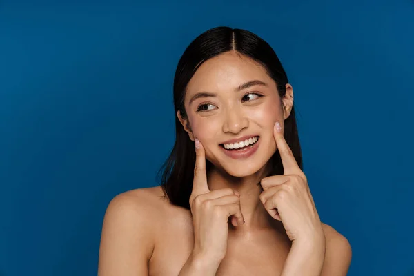 stock image Young beautiful smiling happy asian woman touching her cheeks and looking atside , while standing over isolated blue background