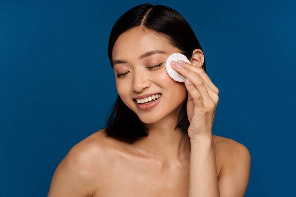 Портрет молодой красивой улыбающейся азиатской женщины с закрытыми глазами, вытирающей лицо хлопковой планкой, стоя на изолированном синем фоне