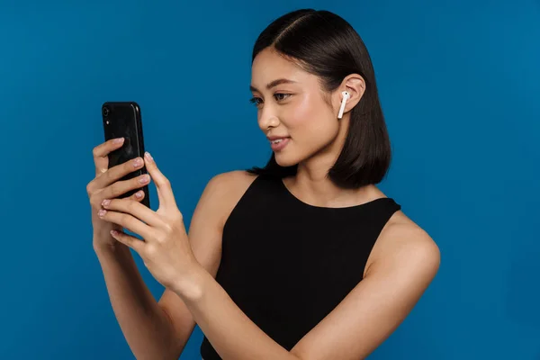 青い背景に隔離された携帯電話を使用している間に笑みを浮かべてイヤフォンのアジアの若い女性 — ストック写真