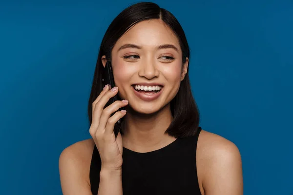亚洲年轻女子一边笑一边用蓝色背景的手机交谈 — 图库照片