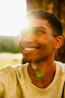 Neşeli, yakışıklı, gündelik giysili Afrikalı bir adam güneşli yeşil parkta otururken yana bakıyor.