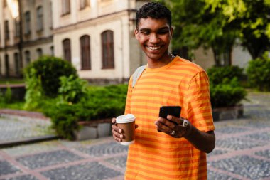 Sıradan giyinmiş gülümseyen Afrikalı genç bir öğrenci üniversite kampüsünde dışarıda dururken akıllı telefon kullanıyor ve kahve içiyor.