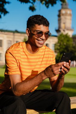 Serbest giyinen neşeli Afrikalı genç bir öğrenci üniversitenin kampüsünde bankta otururken akıllı telefon kullanıyor.