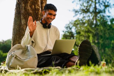 Parktaki ağacın altında otururken gülümseyen ve dizüstü bilgisayar kullanan genç bir zenci.
