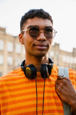 Genç siyah esmer adam piercing takıyor ve güneş gözlüğü takıyor açık hava üniversitesinin önünden geçiyor.