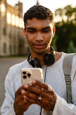 Kulaklıklı ve sırt çantalı genç siyahi adam üniversitenin önünden geçerken cep telefonu kullanıyor.