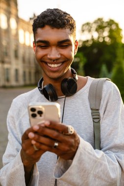 Kulaklıklı ve sırt çantalı genç siyahi adam üniversitenin önünden geçerken cep telefonu kullanıyor.