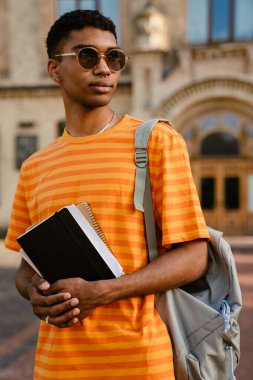 Dışarıda yürürken güneş gözlüğü takan esmer genç bir adam not defteri tutuyor.