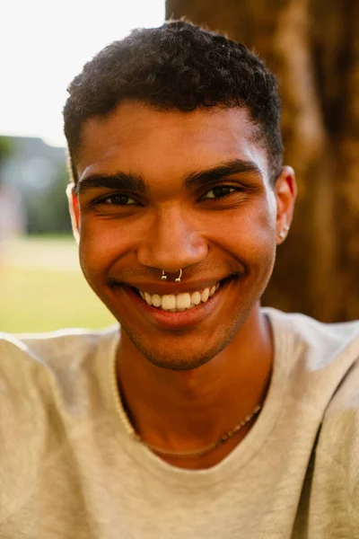 カジュアルな若いハンサムなアフリカ人男性は 晴れた緑の公園で屋外に座ってカメラで笑顔を着ています — ストック写真