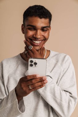 Genç yakışıklı, stil sahibi gülümseyen Afrikalı adam telefona bakıyor ve çenesini tutuyor, izole edilmiş bej arka planda duruyor.