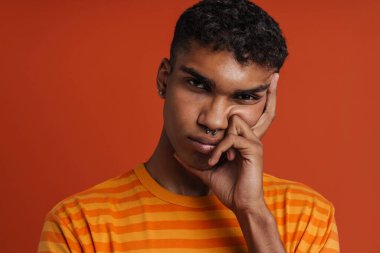 Genç, yakışıklı, ciddi bir Afrikalı adamın portresi yüzüne dokunuyor ve izole turuncu arka planda dikilirken kameraya bakıyor.