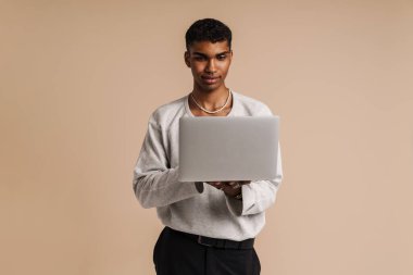 Genç siyah adam bej arka planda kameraya poz verirken dizüstü bilgisayarla çalışıyor.