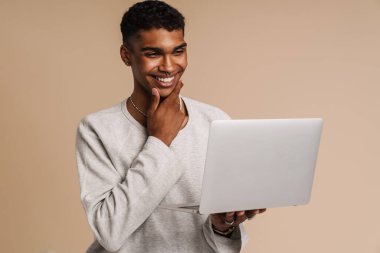 Bilgisayarla çalışırken gülümseyen genç siyah adam bej arka planda izole edilmiş.