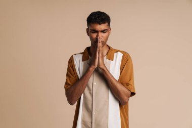 Piercingli genç bir adam bej arka planda izole edilmiş dua jestleri yapıyor.