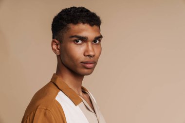 Çekilmiş bej arka planda dikilirken kameraya bakan yakışıklı, ciddi Afrikalı bir adamın profil portresi.