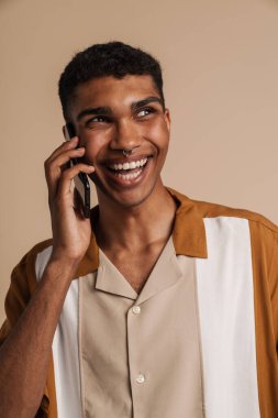 Genç Afrikalı yakışıklı, gülümseyen bir adam telefonla konuşuyor ve bir kenara bakıyor, izole edilmiş bej arka planda duruyor.