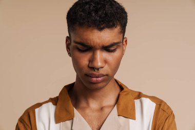 Genç, yakışıklı, üzgün Afrikalı bir adamın portresi. Gözü kapalı ve başı eğik bir şekilde izole edilmiş bej arka planda duruyor.