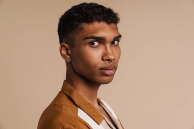 Kameraya piercing takmış yakışıklı, ciddi Afrikalı bir adamın portresi. İzole edilmiş bej arka planda yan yana dururken kameraya bakıyor.